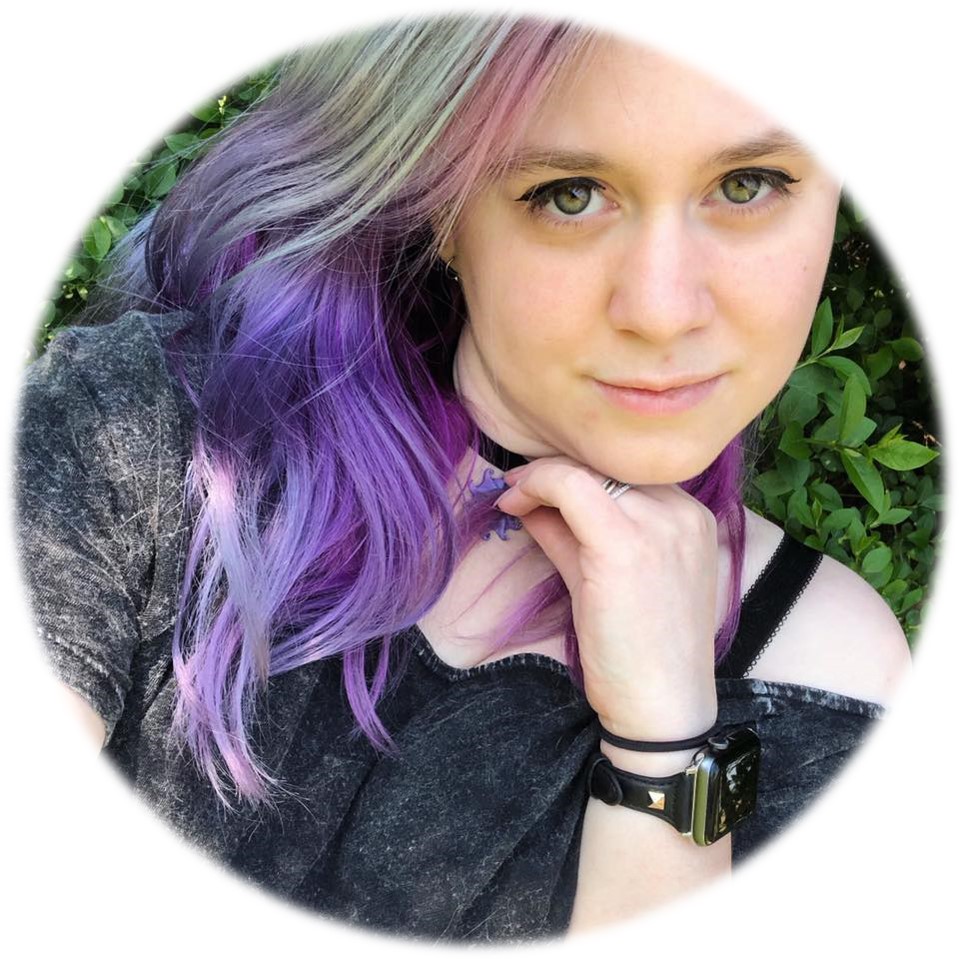 A close up photo of Sarah facing the camera. She is white with purple and blue hair. She is pictured facing the camera with one hand touching her face.